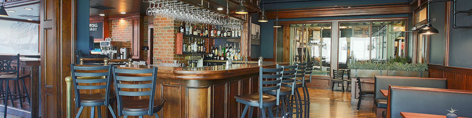 The image shows an empty bar interior with wooden decor, high stools, a well-stocked counter, and booths along the right side.