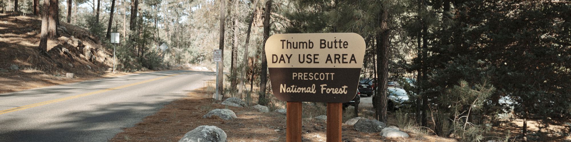 A sign reads "Thumb Butte Day Use Area, Prescott National Forest" next to a road in a wooded area with tall trees and boulders on the ground.