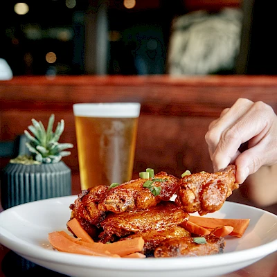 A hand grabbing sauced chicken wings from a plate with carrots, beside a beer and a small plant on a restaurant table.