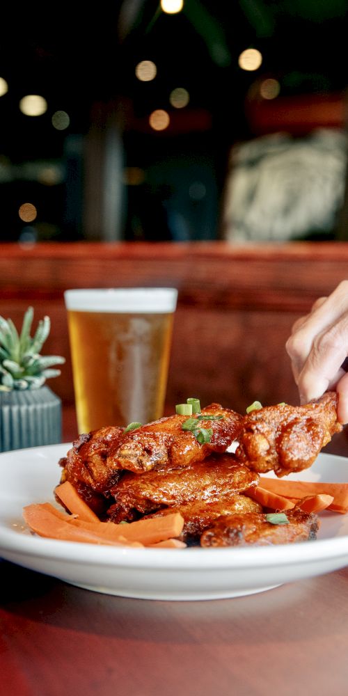 A hand grabbing sauced chicken wings from a plate with carrots, beside a beer and a small plant on a restaurant table.