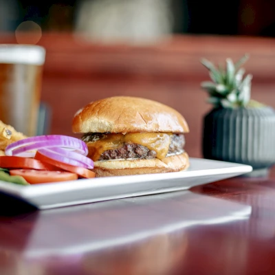 A cheeseburger with fries and vegetables on a plate, a glass of beer, and a small plant in the background on a wooden table.