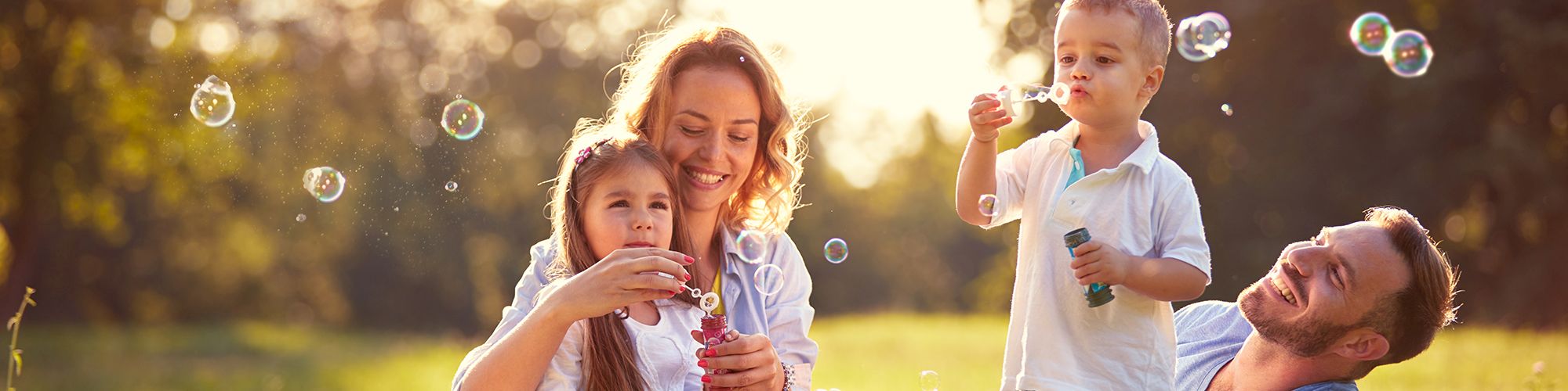 A family of four is enjoying a sunny day in a field, blowing bubbles and smiling together while sitting on the grass.