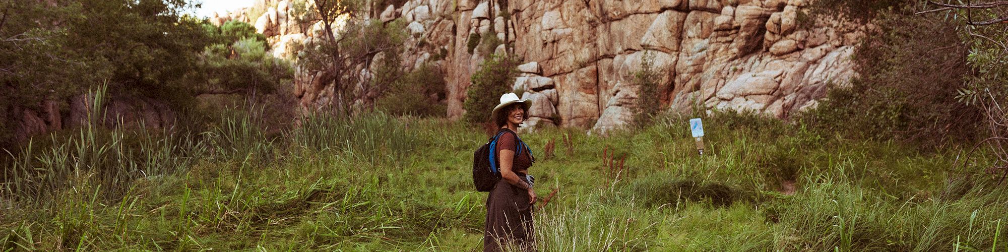 A person wearing a hat and backpack stands in a grassy area surrounded by rocky cliffs and greenery under a clear sky.