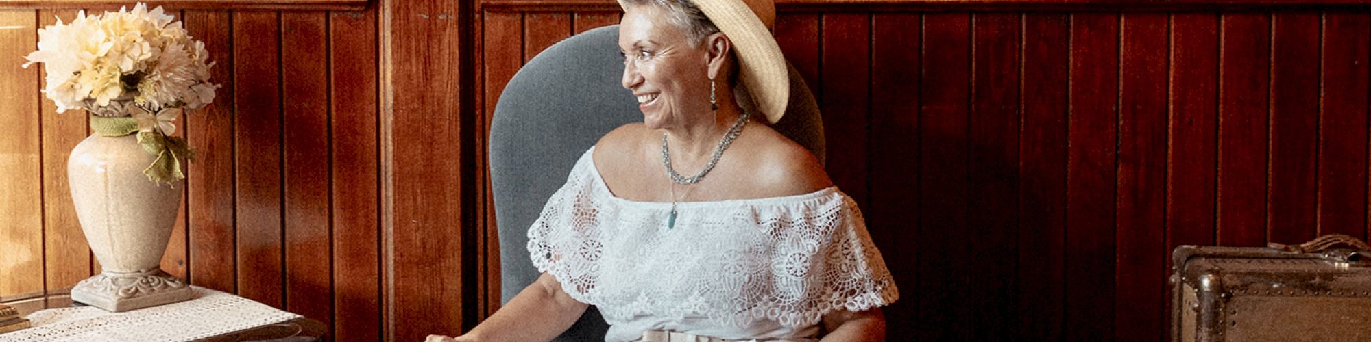 A person wearing a hat sits in a chair in a cozy room with wood paneling, a table with a lamp and flowers, and framed pictures.